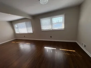 An empty sunlit room with dark hardwood floors and white blinds